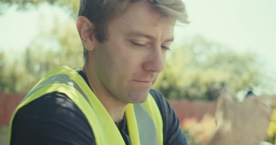 A man in a safety vest sits outdoors on a sunny day holding a walkietalkie to his mouth in a backyard setting