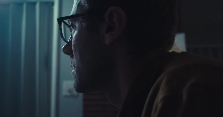A man wearing glasses works on a computer displaying financial graphs at a desk in a dimly lit room