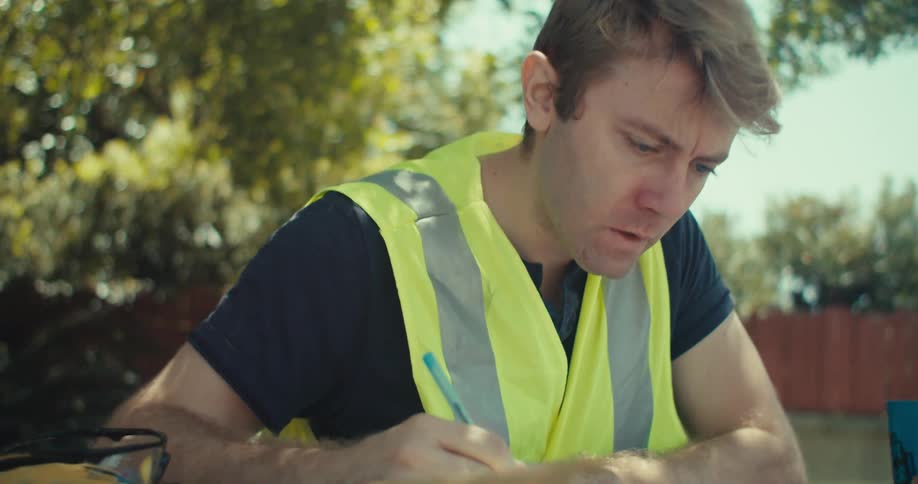A man sitting outdoors at a table writing on a piece of paper with a pen