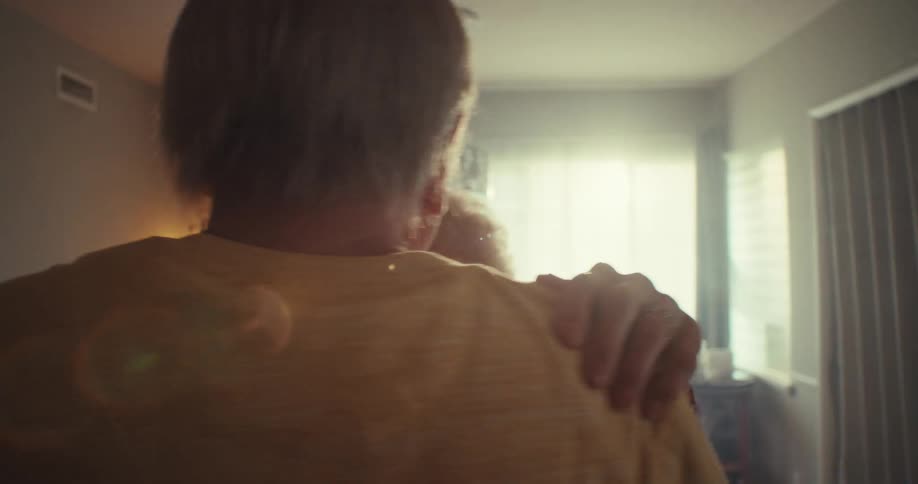 An elderly couple lovingly dances hand in hand in a dimly lit room