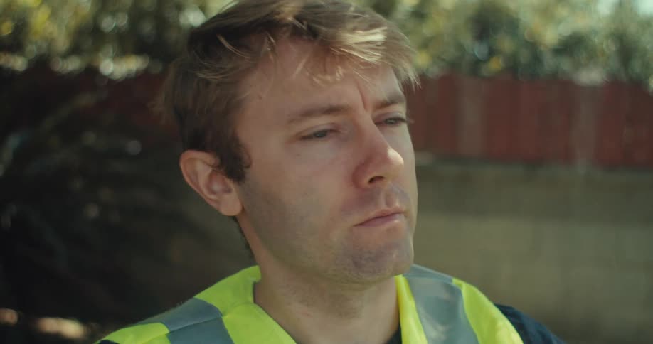 A young man wearing a construction vest sits behind a desk set up on a lawn at midday the desk holds various construction items including a helmet and a measuring tape as the man talks on a walkietalkie