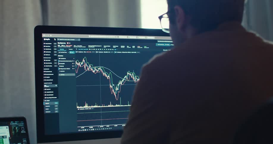 Man seen from behind analyzing stock charts on computer and laptop in a modern workspace