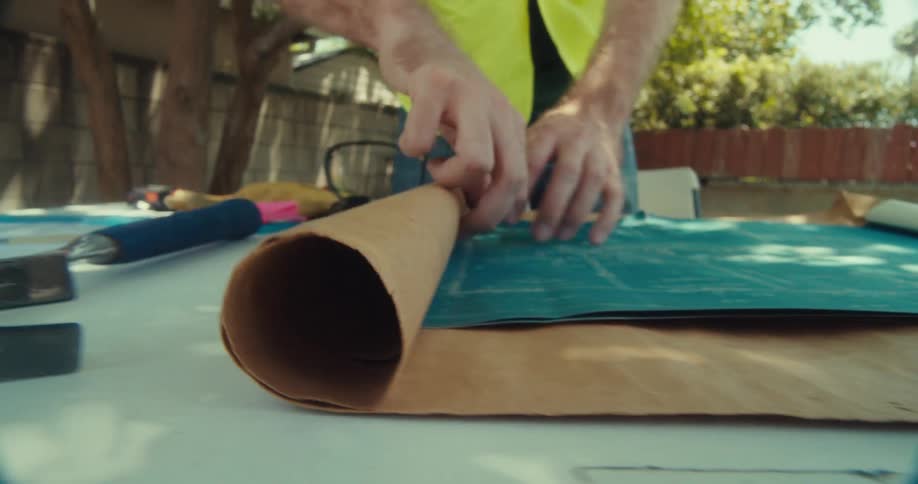 A person in a safety vest is carefully working with blueprints and tools on a table outdoors
