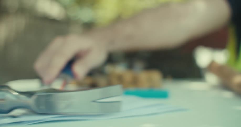 A worker wearing a vest moves a hammer from one side of a table to the other where blueprints are spread out outdoors on a sunny day
