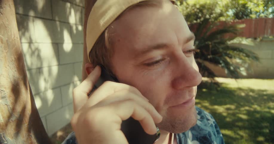 A man sits relaxed under a tree talking on the phone while enjoying a sunny day outdoors
