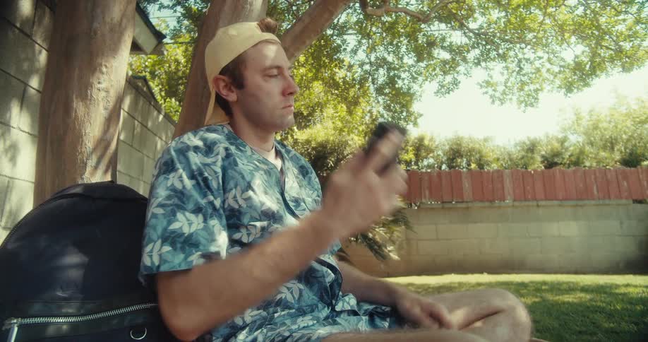 A man wearing a patterned shirt and a baseball cap sits under a tree and talks on a cell phone