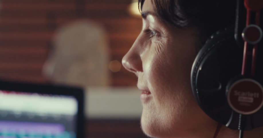 A woman wearing a headset listens and vibes to a song with a computer screen in front of her in a sound studio