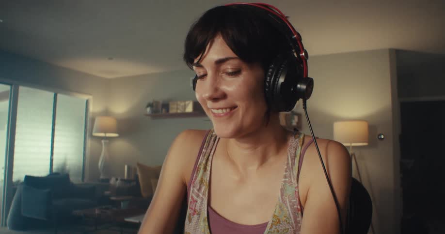 A woman with short dark hair wearing headphones smiles while working on a music keyboard connected to a computer in a living room studio setup