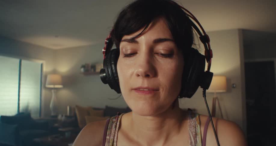 A young woman wearing a headset listening to a song with her eyes closed moving her body to the rhythm in her sitting room at midday