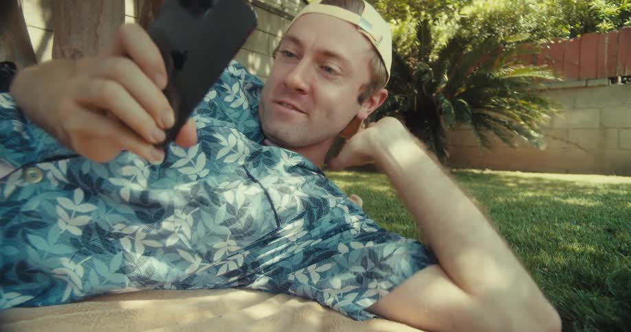 A man lies back in a park smiling while looking at his smartphone