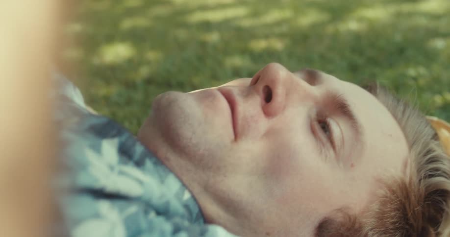 A man lies on the grass in a park smiling while using his smartphone