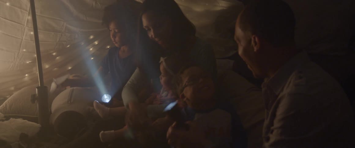 A family sits in a blanket tent with flashlights