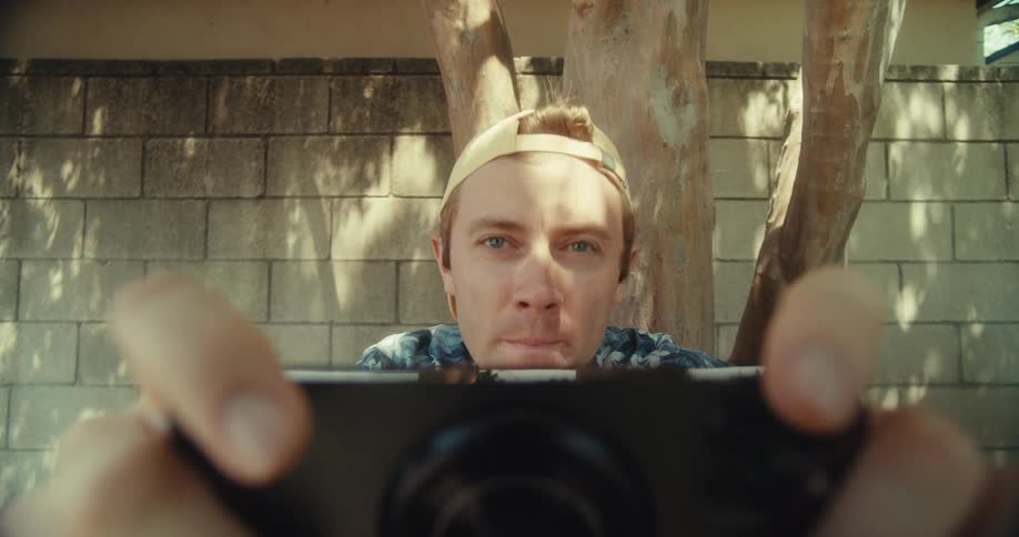 A man sits beside a tree in a yard on a sunny day holding a phone and taking a video shot