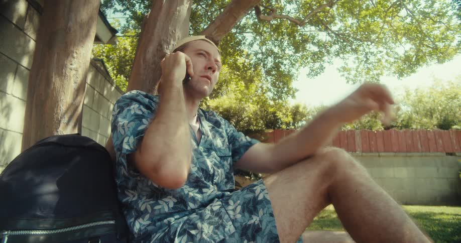 A tourist man sits under a tree engaged in a deep phone conversation
