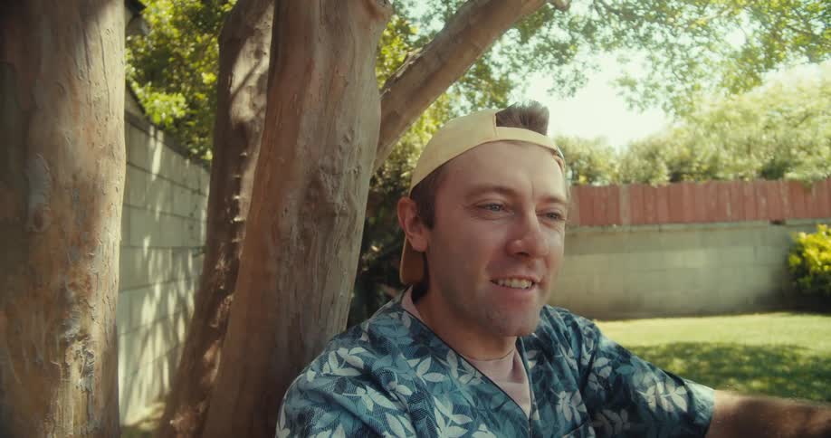A young man takes a selfie with his smartphone while sitting in a backyard on a summer day