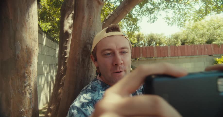 A person sits under a tree in a park happily recording themself and the nature around them