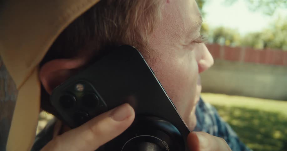 A man in a patterned shirt and a baseball cap sits under a tree picks up his cell phone and starts talking