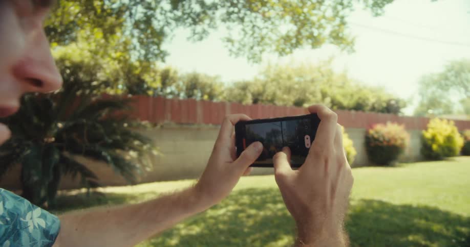 A man leaning against a tree outdoors filming a lawn with his cellphone