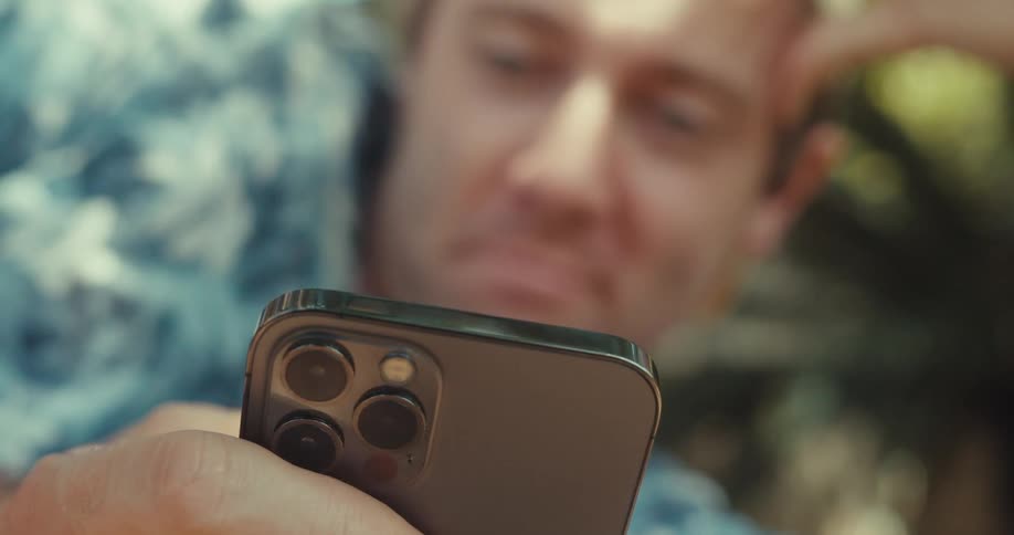 A blurred young man reclined against a backdrop of trees and foliage browsing a smartphone with three rear cameras