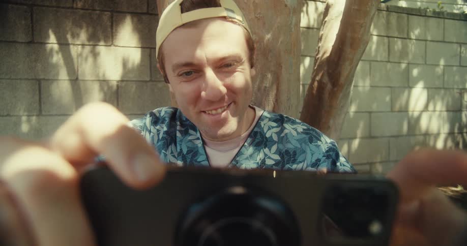 A young man smiles and waves at the camera while filming himself outdoors on a sunny day