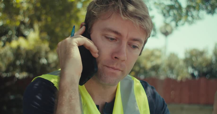 A man in a safety vest talks on a dark cell phone outdoors holding a pen to his ear while reading blueprints