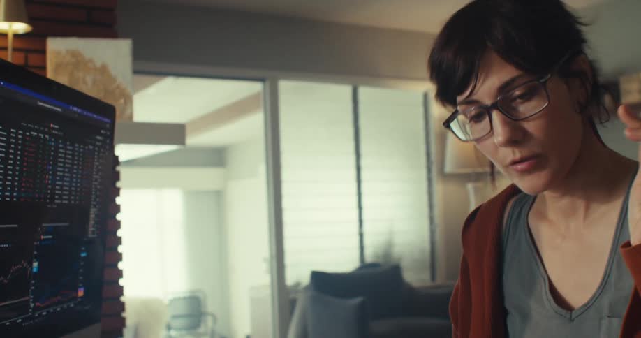 A woman with a ponytail and glasses sits in a room with a brick fireplace looking at a computer screen displaying financial charts