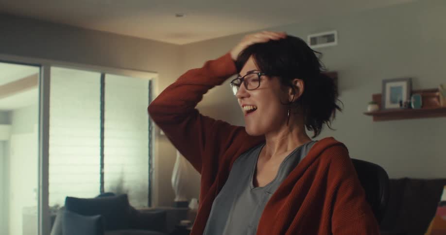 A woman wearing glasses sits in front of a computer in her sitting room laughing with excitement