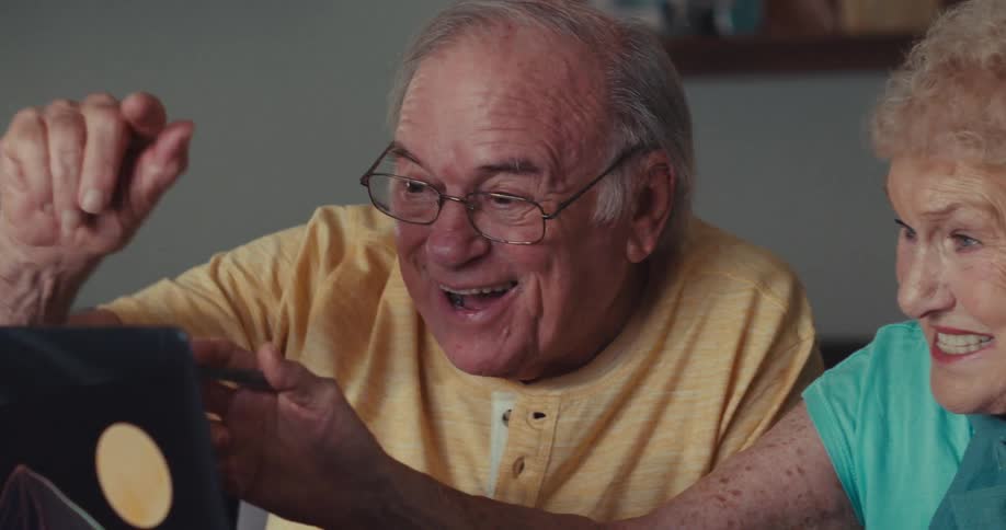 An older man wearing glasses and a yellow shirt points at a laptop screen with a unique mountainandmoon design while laughing and interacting with an older woman beside him who also laughs