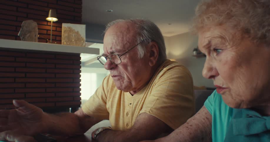 An elderly man wearing glasses and an elderly woman sit side by side in the living room watching a computer screen and talking to each other