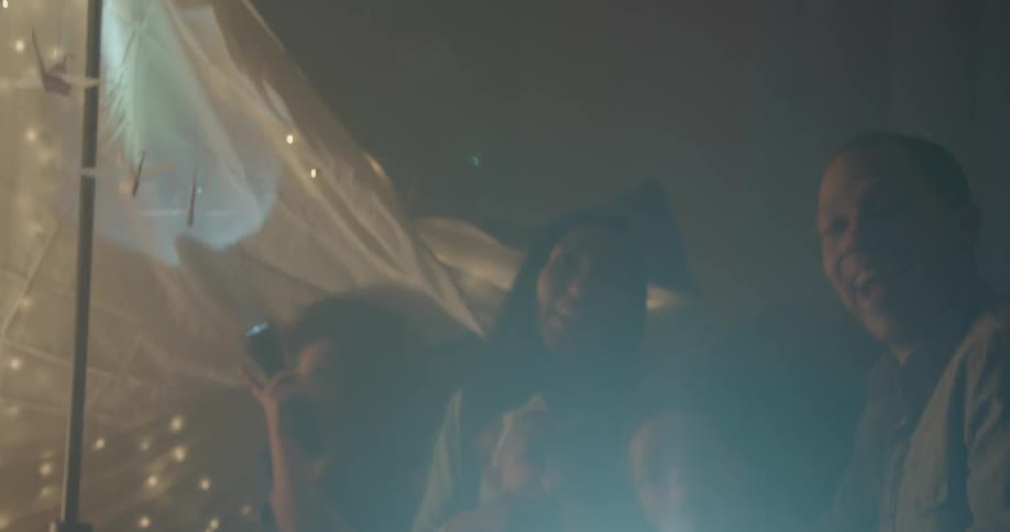 A family sits on the floor near a blanket tent with flashlights