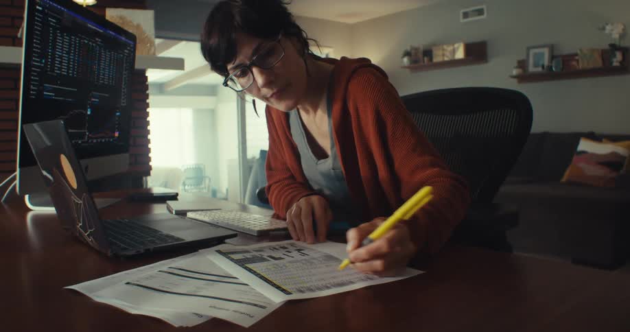 A focused woman is working on a computer and laptop jotting down notes from the screen onto paper