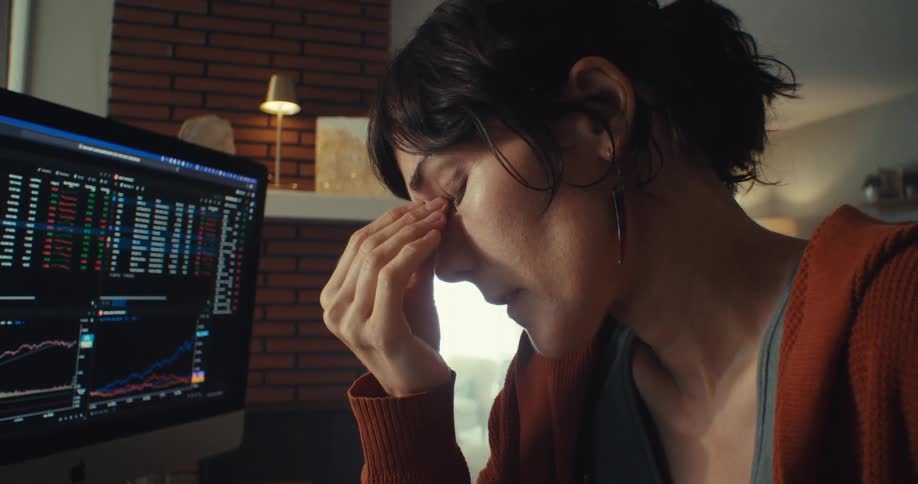 A woman in front of a computer monitor taking off her glasses and placing her hand on her head looking worried and nervous