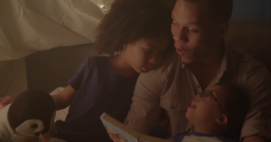 A man reads a book to his children in a blanket tent