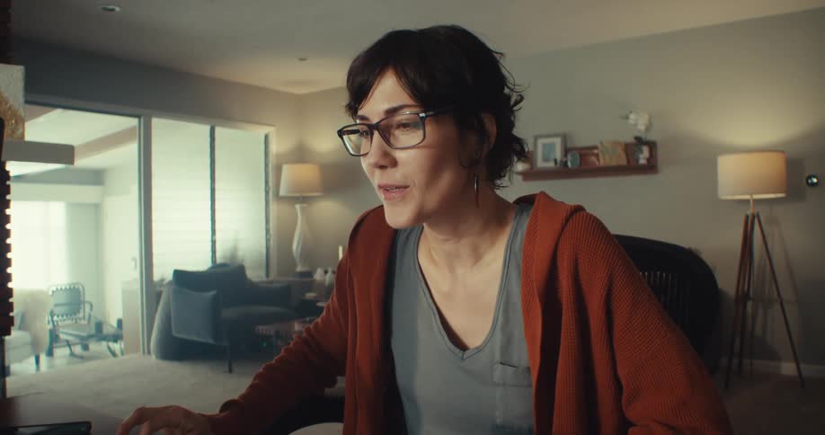 A woman with dark hair and glasses sits at a desk with computer equipment in a living room talking on a video call