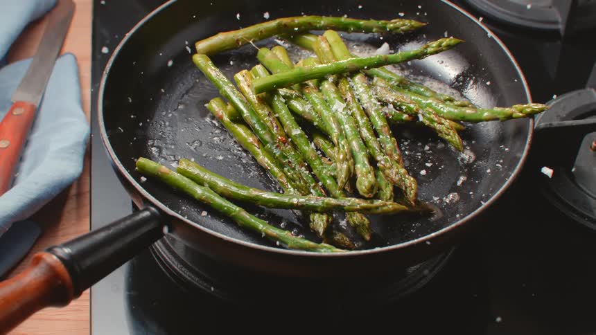 Asparagus and salt flakes fall into a frying pan on a stovetop