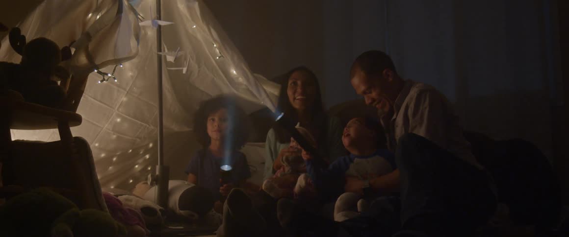 A family sits on the floor with flashlights near a blanket tent