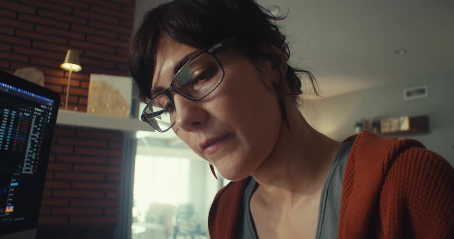 A woman wearing glasses watching a computer screen and jotting down important information with a pen in the living room at midday