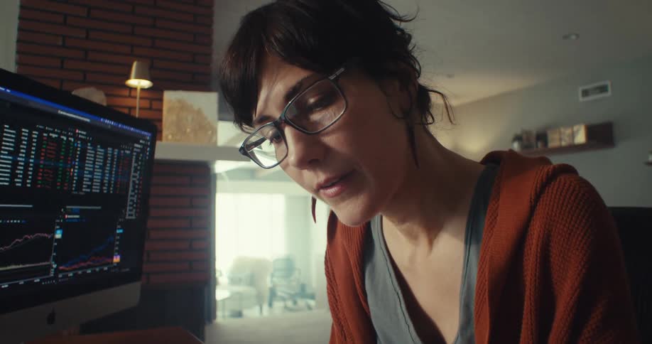 A woman closely reviews financial data on a computer and paper spreadsheets at home