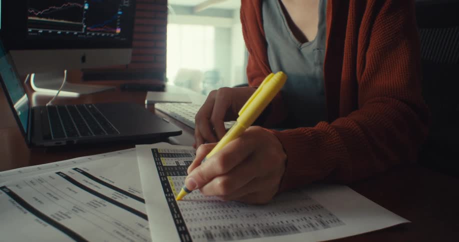 Person highlights printed financial data at desk with laptop and monitor
