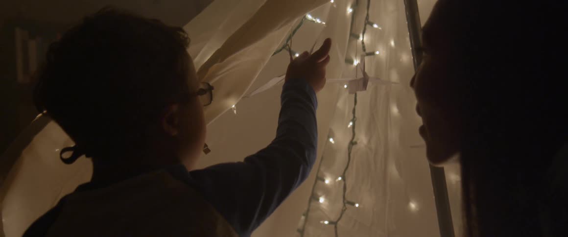 A woman and boy play with origami birds inside a blanket tent