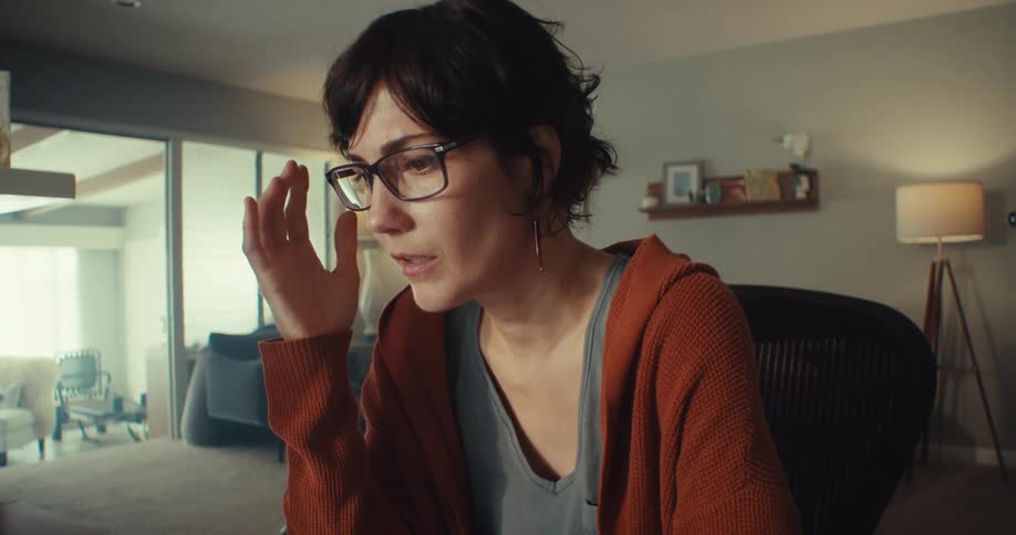 A woman sitting in a living room with her hands on her head looking apprehensive and sad