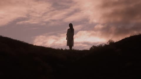 A woman stands alone silhouetted on a hilltop at sunset with dramatic clouds behind her