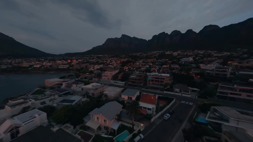 A wide aerial shot shows a coastal neighborhood at dusk with mountains in the background