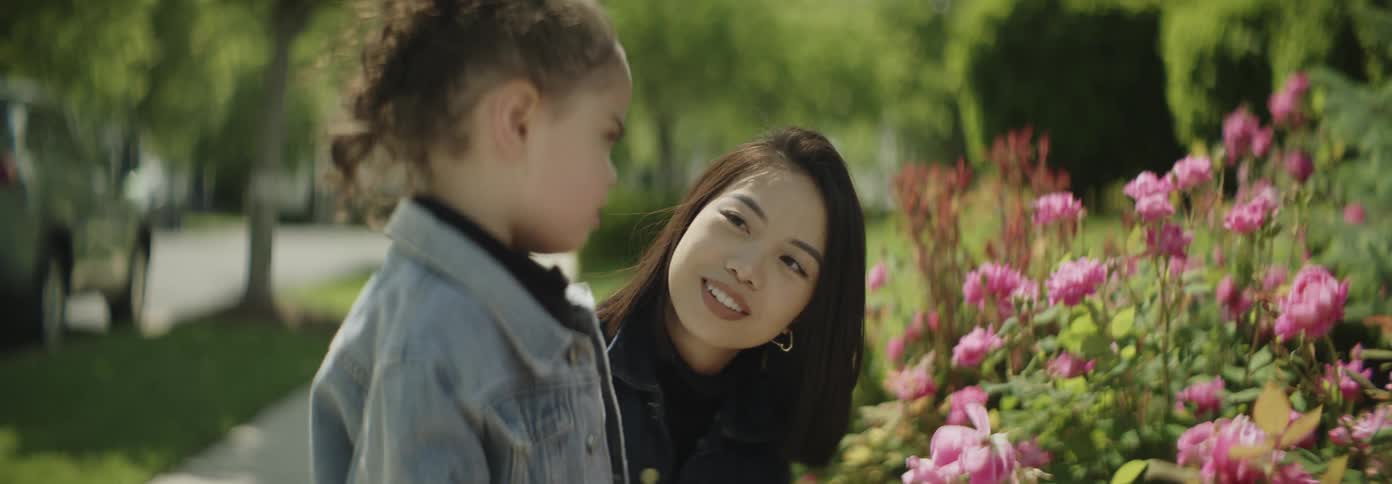 A woman and a young girl stop to smell pink flowers on a sunny day outdoors