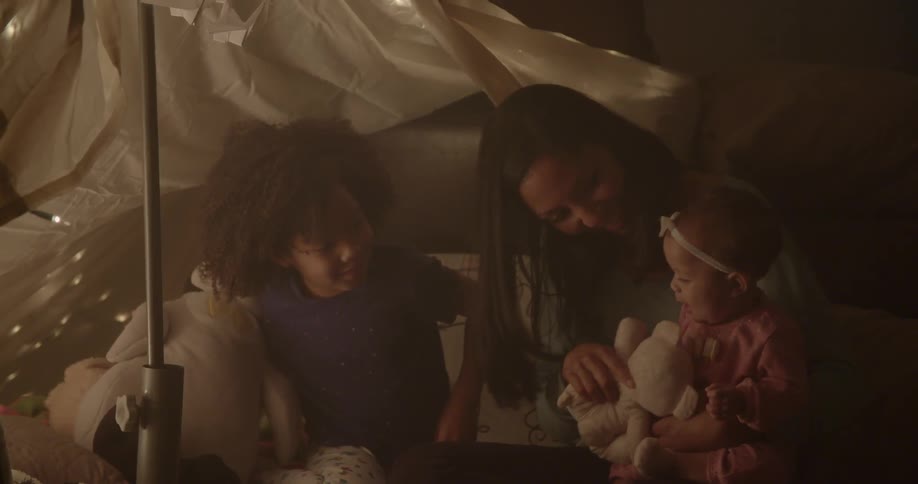 A woman and two children sit under an indoor fort