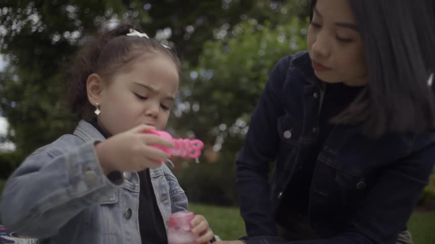 A woman and a child playing outdoors near trees with the child blowing soap bubbles