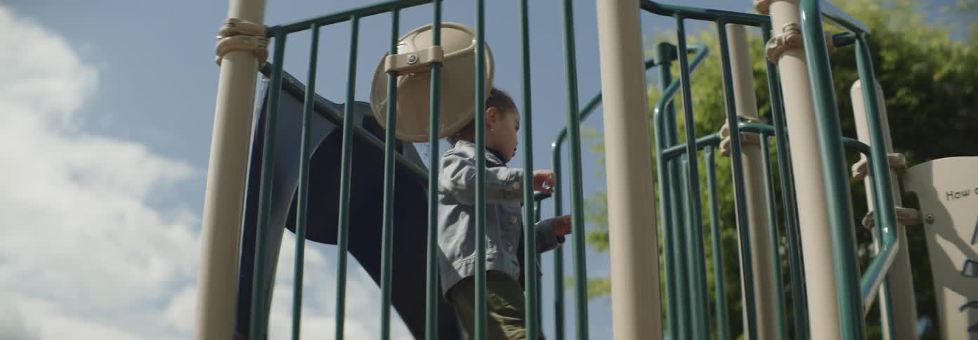 A young child with curly hair climbs up a piece of playground equipment in a park