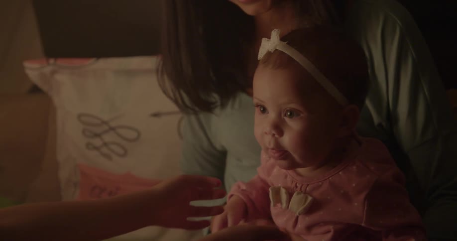 A little girl plays with a baby who is sitting on a woman's lap in a dark room