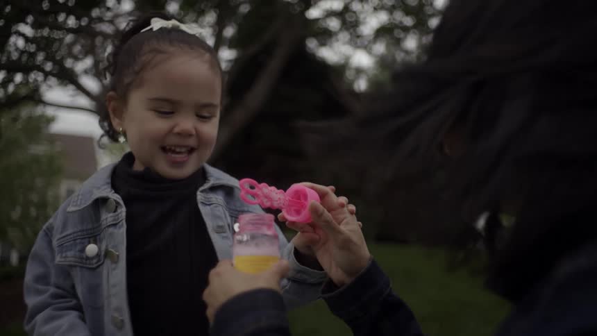 A child laughs and runs after bubbles while her mother blows them holding a bottle of bubble solution outdoors