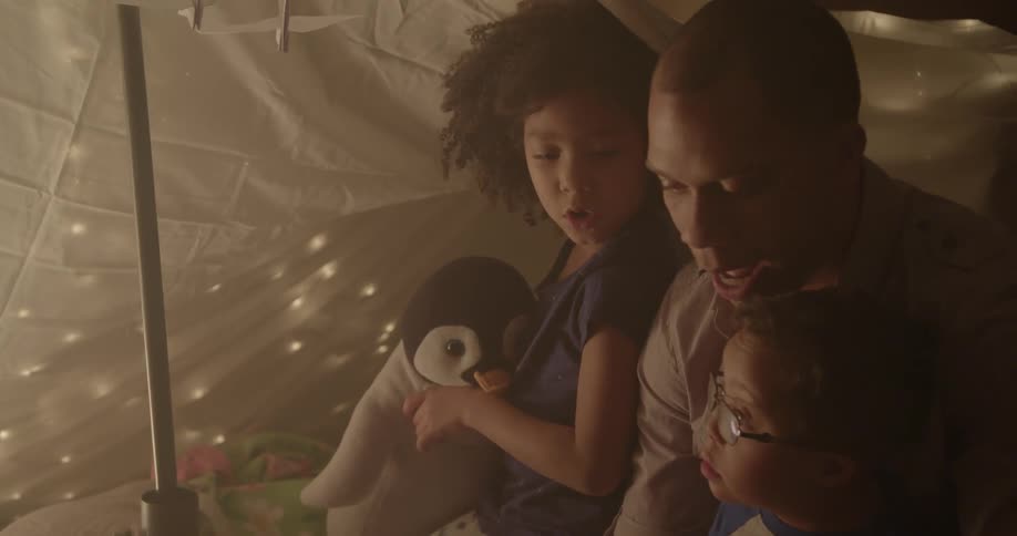 A man and young children sit inside a blanket tent while reading a book